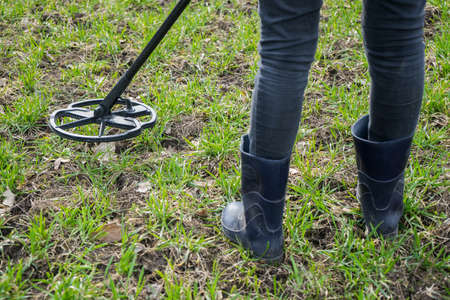 girl archaeologist or metal digger is looking for coins or metal with a metal detector. Search old coins. Scrap metal collection on autumn. Girl in rubber boots with a tool for finding coins. の写真素材