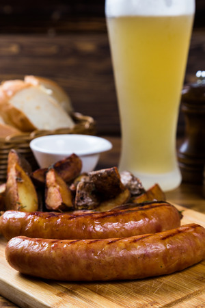 Grilled sausages with roast potato with spices on wooden board with light beer on tableの写真素材