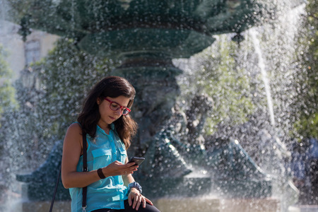 young girl in blue blouse and red glasses with a phone on the near the fountain of European citiesの写真素材