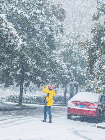 A woman in a yellow jacket and jeans is standing in the middle of a snow-covered road during a heavy snowfall.の写真素材