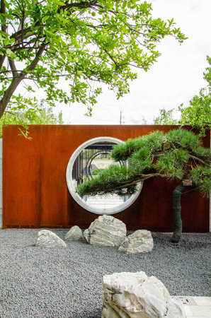 HANGZHOU, CHINA - JULY 03, 2017: Vertical shot of a rusty Corten steel wall with a circular opening (moon gate) in a Zen-style garden. White rocks and gray gravel contrast with the industrial metal texture and green trees.のeditorial素材