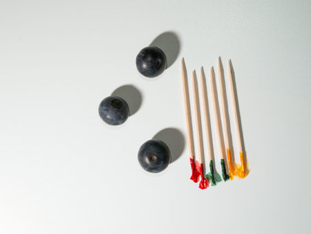 A minimalist layout featuring three fresh blueberries arranged next to five bamboo cocktail toothpicks with colorful frilled ends on a plain white surface.の写真素材