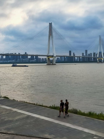 Wuhan, Hubei Province, China - May 30, 2023: People walk along the concrete riverfront pathway with...
