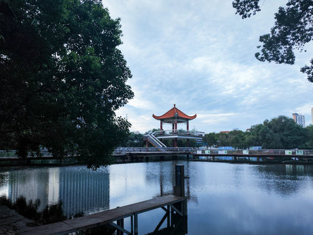 A wide perspective of a traditional red-roofed pavilion standing on a bridge over a calm lake,...