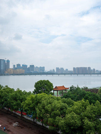 A wide-angle landscape photograph featuring a modern cityscape skyline across a body of water, with a lush green park and a traditional Chinese-style pavilion in the foreground. The scene represents urban development, city planning, and recreational public spaces in a harmonious setting.の写真素材