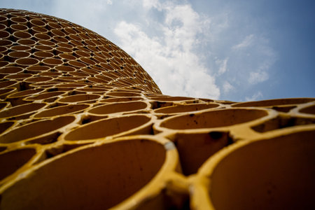 A low-angle perspective looking up at a golden honeycomb-patterned metal sculpture against a cloudy...