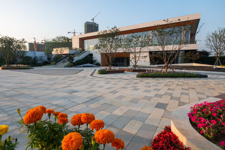 A sunny plaza featuring a modern building, paved ground, and bright orange flowers in the foreground. The image conveys urban design, color, and outdoor leisure. High-quality photography.の写真素材