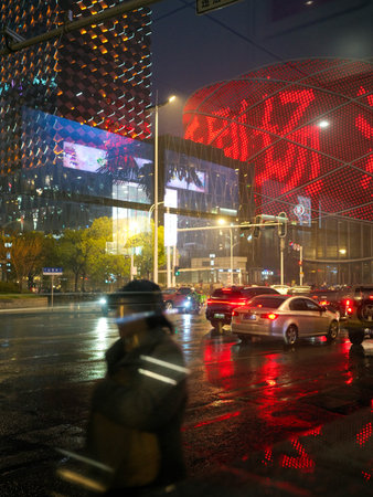 Wuhan, China - March 12, 2025: Silhouetted pedestrian walking past illuminated commercial buildings at night. The image shows rainy weather and city light reflections on the road.のeditorial素材