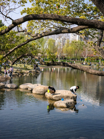 Wuhan, China - March 22, 2025: A group of people catching small fish or tadpoles on large stepping stones in a reflective park pond. The image shows the water surface, overhanging trees, and distant buildings.のeditorial素材