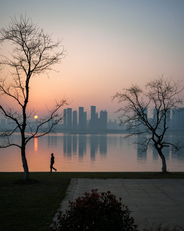 Wuhan, China - March 23, 2025: A person walking past a silhouetted tree at the waterfront during sunset. The image shows the sun reflecting on the water and a hazy city skyline.のeditorial素材