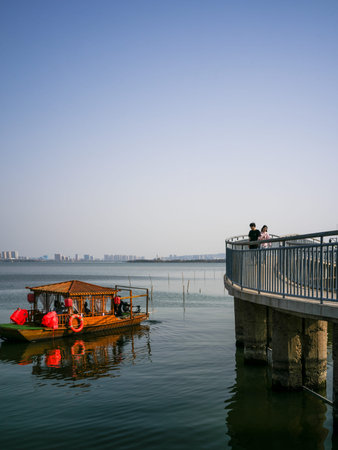 Wuhan, China - March 24, 2025: Wooden tourist boat sailing on the water near a curved observation deck by a lake. The image shows people standing by a metal railing, concrete pillars in the water and a distant city skyline.のeditorial素材