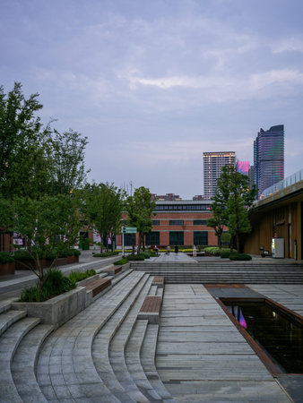 Wuhan, China - June 11, 2025: Wide urban public square featuring curved stone steps, wooden decking and a reflection pool at dusk. The image shows red brick cultural buildings, trees and distant illuminated high-rise architecture.のeditorial素材