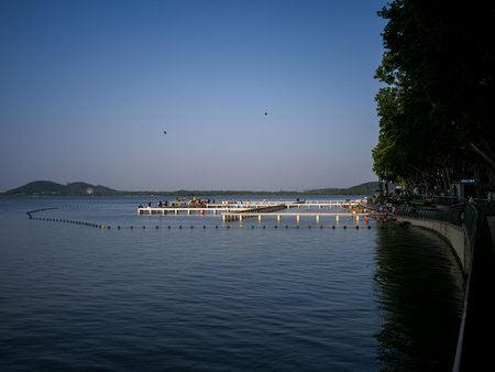 Wuhan, China - June 16, 2025: Distant view of people walking on a long white pier extending into the calm waters of a lake. The image features buoys marking a swimming area, large trees on the shore and hills in the distance.のeditorial素材