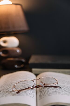 Still life with old books and reading glasses lit by night light. Calm evening atmosphere in a dark brown room.の写真素材