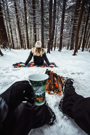 Young romantic couple having fun outdoors in the forest in winter season. Pov view of man near the campfire and holding hot tea cup. .の写真素材