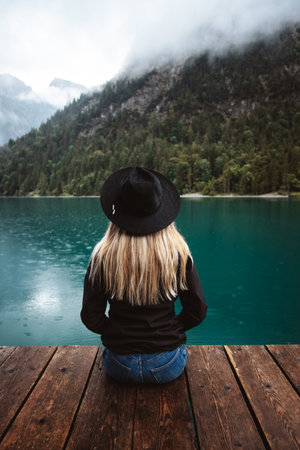 Woman sitting on a wooden pier looking at a lake in the mountainsの写真素材