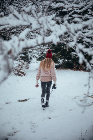A traveler girl in a pink jacket and a red hat walks through the snowy forest with a camera in...