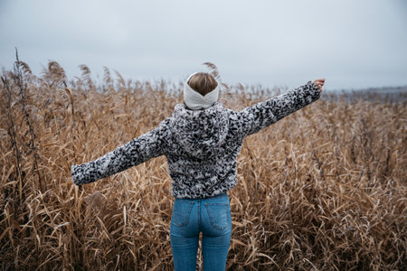 A girl in a knitted sweater and jeans stands on the edge of a dry reed field.の写真素材