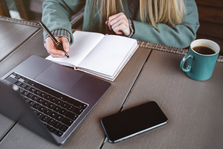 Female entrepreneur sitting at a table in home terrace of a coffe shop and writing down notes and...