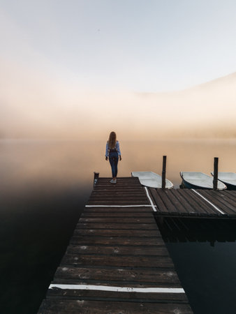 Young woman standing on wooden jetty at foggy lake in morningの写真素材
