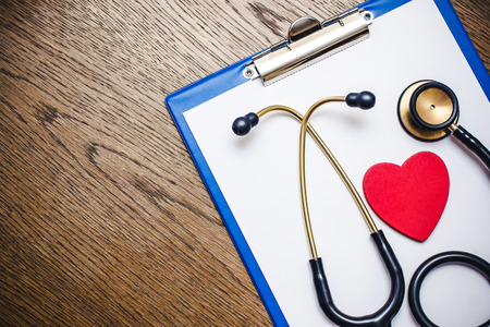 Doctor clipboard stethoscope and heart shape on white paper. Brown wooden table.の写真素材