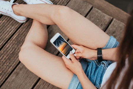 Girl sitting on wooden floor using mobile phone. Watching summer exotic sunset landscape on...