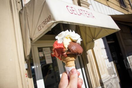 Ice cream summer vacation in Italy. Gelateria shop in the background and hand holding waffle cone with three types of ice cream.のeditorial素材