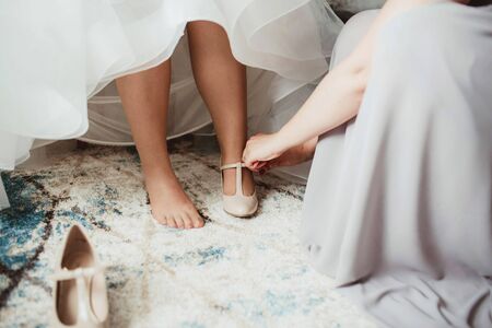 Young bride getting ready for ceremony. Dressing up white wedding dress. Wearing shoes preparation background. Bridesmaid help and support.の写真素材