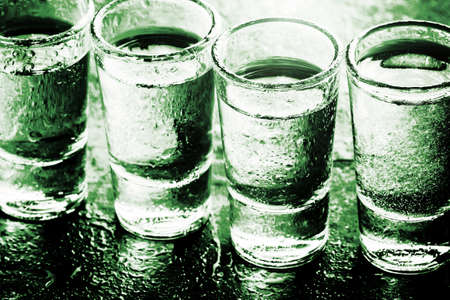 Glass vodka shots. Many alcohol drinks in a row texture. Weekend party background. Green wet bar surface reflection. Liquor drops spilled. Backlight transparent glass backdrop.の写真素材