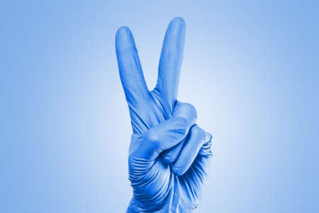 Hand in blue latex glove shows victory gesture on a blue background.の写真素材