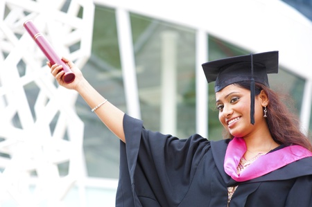 Happy young Indian in a graduation gown and cap holding her diplomaの写真素材