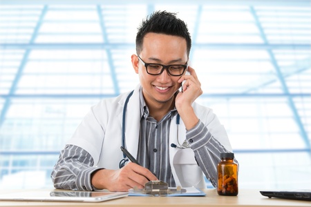 Young Southeast Asian medical doctor writing a note while calling on the phone, sitting in front a desk inside hospital.の写真素材