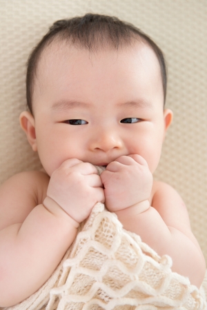 Adorable six months old teething Asian baby girl lying on bed biting blanket.の写真素材
