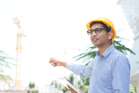 Portrait of a smiling Indian male contractor engineer with hard hat pointing to a construction siteの写真素材