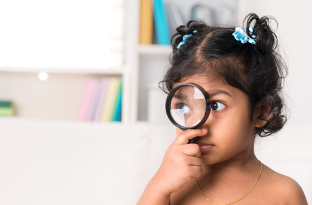 Child at home. Cute Indian girl peeking through magnifying glass.の写真素材