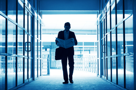 Silhouette of business man residing newspaper and walking thru modern office building, in blue tone.の写真素材