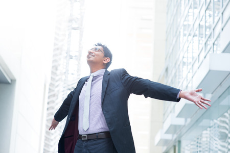 Indian businessman open his arms wide, looking up into the light, modern office building as background, natural sunlight.の写真素材