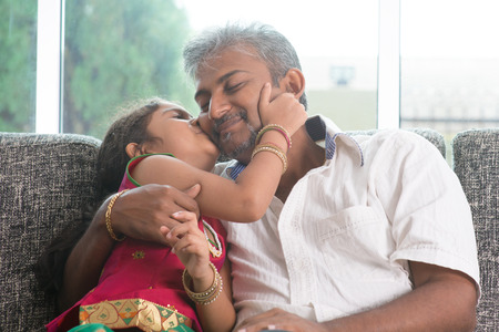 Happy Indian family at home. Asian sweet daughter kissing her father face indoor, sitting on sofa.の写真素材