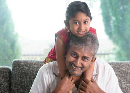 Happy Indian family at home. Asian father piggyback her daughter, sitting on sofa. Parent and child indoor lifestyle.の写真素材