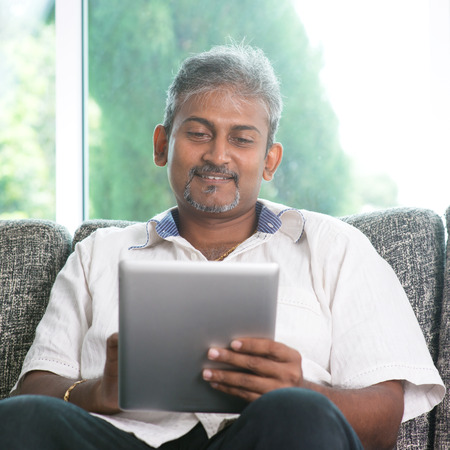 Portrait of middle aged Indian man reading on digital tablet computer and smiling at home.の写真素材