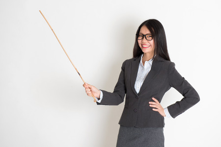 Asian female teacher smiling and holding a stick, standing on plain background.の写真素材