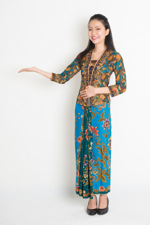 Full length portrait of Southeast Asian woman in batik dress hand holding something standing on plain background.の写真素材
