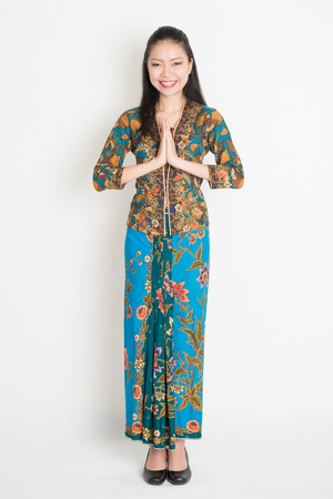 Full length Southeast Asian woman with batik dress in greeting gesture standing on plain background.の写真素材