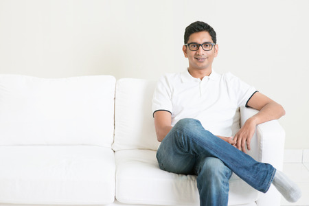 Good looking young Indian guy sitting on sofa and smiling. Lifestyle Asian man at home. Handsome male model.の写真素材