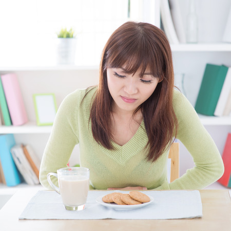 Woman holding stomach and suffering stomachache, while having dairy milk and cookies as breakfast, sitting at dining table.Young people indoors living lifestyle at home. Woman holding stomach and suffering stomachache, while having dairy milk and cookies as breakfast, sitting at dining table.Young people indoors living lifestyle at home.