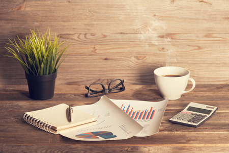 Workplace with booklet, pen, glasses, cup of coffee and graphs. Wooden table background in vintage toned.の写真素材