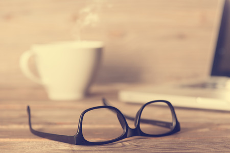 Wooden work table with glasses, laptop, cup of hot coffee, in dramatic light vintage toned.