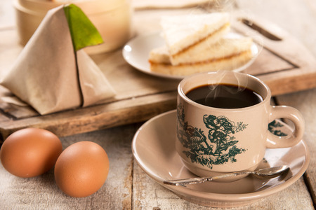 Traditional kopitiam style Malaysian coffee in vintage mug and saucer and breakfast with morning sunlight. Fractal on the cup is generic print. Soft focus dramatic ambient light over wood table.の写真素材