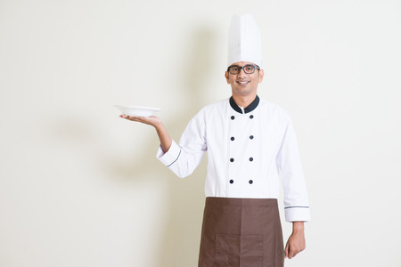 Portrait of handsome Indian male chef in uniform presenting an empty plate and smiling, standing on plain background with shadow, copy space.の写真素材