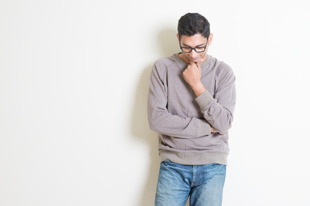 Portrait of handsome casual Indian male thinking and looking downwards, standing on plain background...